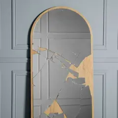 A tall, arched mirror with a wooden frame is shattered, with many glass shards reflecting a grey, paneled wall.
