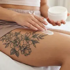 Eine Frau pflegt ihr **oberschenkel tattoo klein** mit einer Creme. Das filigrane Blumenmotiv ziert ihren Oberschenkel.