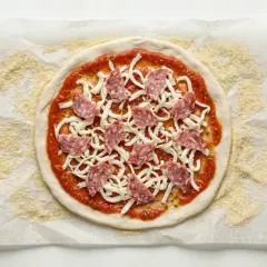Okrągłe ciasto na pizzę z sosem, serem i salami, przygotowane na papierze do pieczenia.