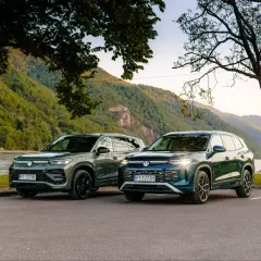 Dwa nowe VW Tayron, zielony i granatowy, zaparkowane nad rzeką, z górami w tle.