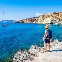 Un voyageur admire la mer Égée bleue et les falaises blanches, un voilier au loin. Le tourisme en Grèce offre des paysages magnifiques.