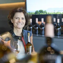 Frau mit Weinflasche und Glas, im Hintergrund Flaschenreihe und Landschaft.