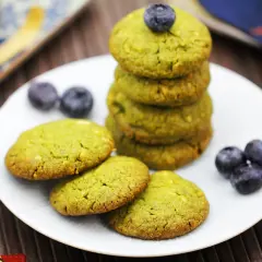 Grüne matcha kekse mit Blaubeeren auf einem Teller, daneben ein Matcha-Schneebesen.