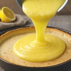 Une crème onctueuse au lemon curd est versée dans une tartelette dorée. Des citrons coupés en deux sont en arrière-plan.