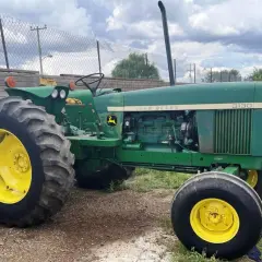 Zielony traktor John Deere 3130 z żółtymi felgami stoi na żwirowym podwórzu, gotowy do pracy na polu.
