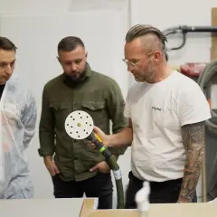Trzech mężczyzn przygląda się polerce. Jeden z nich, w białym kombinezonie, wygląda na ucznia. To szkolenie z obróbki kamienia, jak w grze **stone age**.