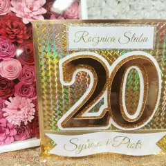 Złota kartka z napisem "Rocznica Ślubu" i liczbą "20" dla Sylwii i Piotra. Obok ramka z różowymi papierowymi kwiatami.