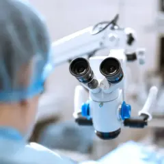 Mikroskop chirurgiczny w rękach lekarza w czepku i fartuchu, w tle sprzęt medyczny.