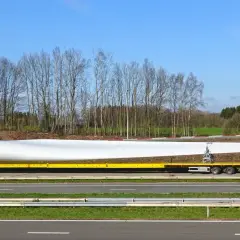 Żółta ciężarówka transportuje ogromne łopaty wiatraka. Ten pojazd nienormatywny pokonuje drogę, by dostarczyć element do budowy farmy wiatrowej.