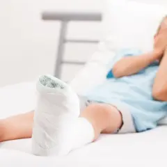 Chłopiec z gipsie na nodze, po złamaniu kości piszczelowej, zakrywa twarz dłońmi.