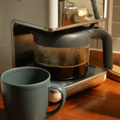 En kaffebryggare med en glaskanna full med färskt kaffe. Bredvid står en mugg och en stapel tallrikar. Man kan brygga presskaffe i kaffebryggare för en god start på dagen.