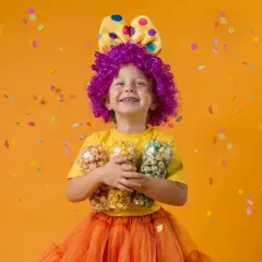 Niña con disfraz casero de payaso, peluca morada, lazo amarillo con lunares y falda naranja, sosteniendo tres vasos de palomitas de colores.