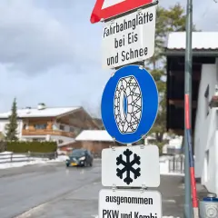 Verkehrsschilder weisen auf Fahrbahnvereisung bei Eis und Schnee hin. Ein blaues Schild zeigt eine Schneekette, ein weiteres ein Schneeflockensymbol.
