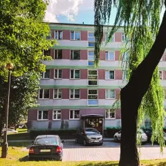 Blok mieszkalny z zielonymi i różowymi pasami, drzewa i zaparkowane samochody.