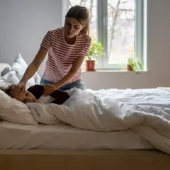 Kobieta sprawdza czoło śpiącego dziecka, które leży w łóżku pod kołdrą. Wygląda na to, że dziecko ma przeziębienie.