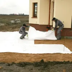 Dwóch mężczyzn rozkłada białą geowłókninę, przygotowując tani taras.