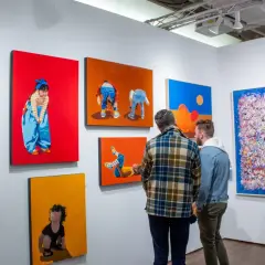 Two men examine colorful paintings at an art fair, perhaps contemplating their next acquisition as an art collector.