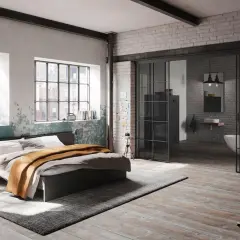 Ein gemütliches **Bett unter Fenster** mit rustikalem Charme. Daneben ein offener Bereich mit Badewanne und Waschbecken.