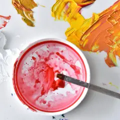 Ein Pinsel taucht in rote Farbe. Daneben sind gelbe und orangefarbene Farbkleckse. So mischt man rot.