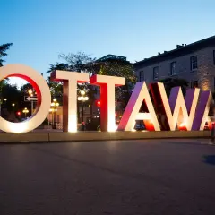 Napis OTTAWA, stolica Kanady, świeci w nocy. W tle budynki i drzewa ozdobione lampkami.