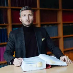 Mężczyzna w garniturze siedzi przy stole z otwartą książką, w tle regały z książkami.