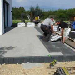 Pracownicy układają płyty na tarasie betonowym. W tle koparka i maszyna budowlana.