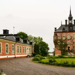 Vackert vik slott med en lång, röd tegelbyggnad och ett torn med klocka. Grönskande trädgård och en grusväg leder fram till byggnaderna.
