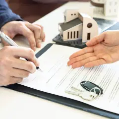 Signature d'un contrat immobilier. Un homme signe un document, des clés et un modèle de maison sont posés sur la table. Le courrier sru est prêt.
