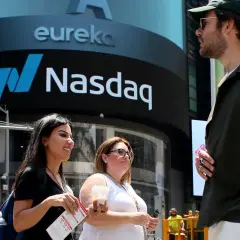Ludzie przed budynkiem Nasdaq, gdzie handluje się akcjami. Słońce świeci, tworząc cień.
