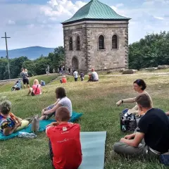Ludzie odpoczywają na trawie pod kamienną wieżą i krzyżem. Trasa na Święty Krzyż przyciąga turystów.