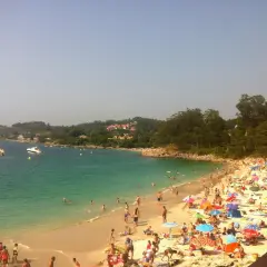 Gente disfrutando del sol y el mar en la playa areacova, con barcos en el agua y colinas verdes al fondo.