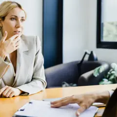 Kobieta w jasnej marynarce zamyślona, ręką dotyka ust. Rozmowa przy biurku, dokumenty. Bank odmówił kredytu po podpisaniu umowy, czeka na wyjaśnienia.