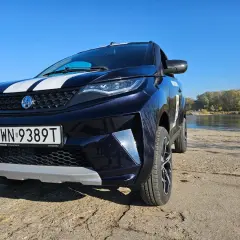 Ciemnoniebieski SUV z białymi pasami na masce, zaparkowany nad rzeką. To auto od 14 lat, gotowe na przygodę.