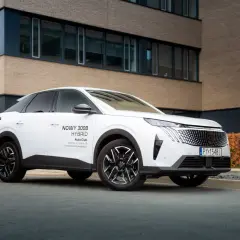 Biały samochód na e, nowy Peugeot 3008 Hybrid, zaparkowany przed nowoczesnym budynkiem z ceglaną elewacją.