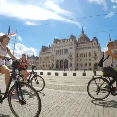 Friends enjoying Budapest bike tours, cycling past the Parliament building under a bright, cloudy sky.