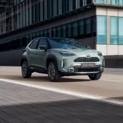 En grön Toyota Yaris Cross hybrid kör på en stadsgata. Bilen är i fokus framför moderna byggnader.
