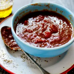 En skål med tjock, röd bbq sås med bitar av tomat. En sked ligger på en tallrik med salt och peppar.