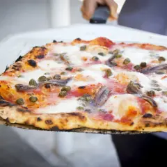 Pizza na redukcji z anchois i kaparami, wypiekana na cienkim cieście, prezentowana na łopacie.