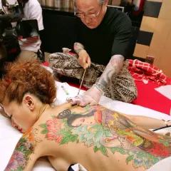 Mistrz tatuażu tworzy tradycyjne tatuaże w Japonii na plecach kobiety. W tle operator kamery dokumentuje proces.