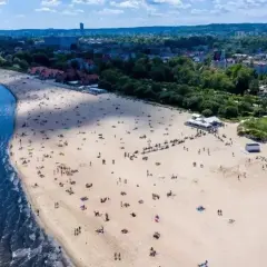 Słoneczny dzień, plaża pełna ludzi, idealne miejsce, gdzie pociągiem nad morze. W tle widać miasto i zieleń.