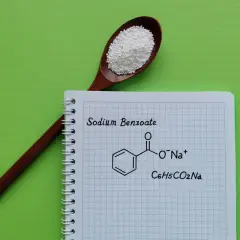 Drewniana łyżka z białym proszkiem, obok notatnik z napisem "Sodium Benzoate" i jego wzorem chemicznym.
