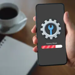 Dłoń trzyma smartfon z ikoną narzędzi i paskiem postępu "Factory Reset". Kawa i notatnik w tle.