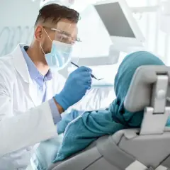 Lekarz stomatolog w masce i fartuchu bada pacjentkę w chuście. Nawet przy dentofobii, troska i profesjonalizm mogą pomóc.