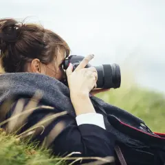 Kobieta z włosami spiętymi w kok leży na trawie, robiąc zdjęcie profesjonalną lustrzanką do vlogów.