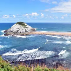 Olas rompen en las **playas limpias de Cantabria**, con un islote rocoso y vegetación verde.