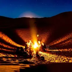 Un groupe se réchauffe autour d'un feu de camp dans le désert du Sahara la nuit. La température chute, mais la chaleur du feu et la beauté du ciel étoilé créent une atmosphère magique.