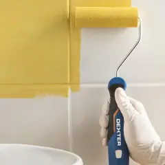 Mano con guante blanco pintando azulejos de baño de blanco a amarillo con rodillo.