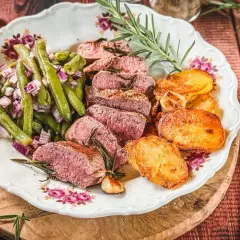 Saftiger Lammbraten nach Omas Rezept, serviert mit grünen Bohnen, roten Zwiebeln und knusprigen Kartoffelscheiben auf einem Teller mit Blumenmuster.