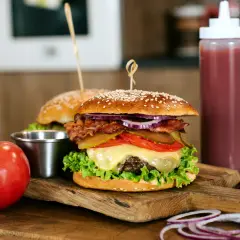 Soczysty burger wołowy z bekonem, serem i warzywami na desce, obok pomidorów i sosu.