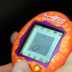 Pomarańczowe Tamagotchi Connection w dłoni, z widocznym ekranem i przyciskami. Pamiętasz, co to tamagotchi?